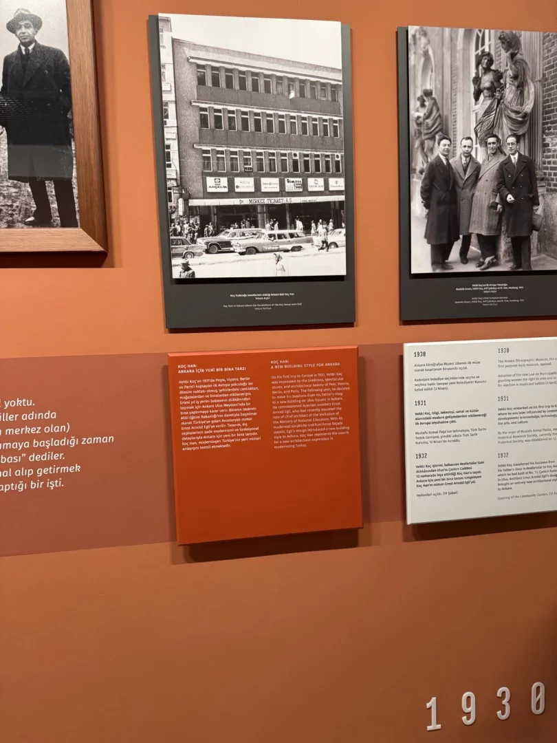 Turuncu bir sergi duvarında asılı, Koç Topluluğu'nun temellerinin atıldığı Ankara'daki çok katlı Koç Hanı'nı gösteren, önünde klasik arabalar ve yayalar bulunan, zemin katında Dr. Merkez Ticaret A.Ş. ve Arçelik gibi mağazaların yer aldığı siyah beyaz bir fotoğraf ve yanında Koç Han'ın yeni bina tarzını anlatan bilgilendirme panelleri,