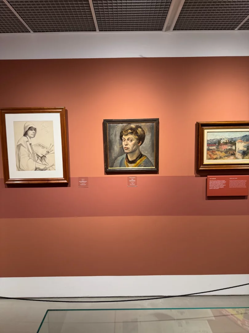 Terracotta renkli duvarda, solda resim malzemeleriyle şapkalı bir figürün eskizi, ortada mavi yakalı sarı bluzlu bir kadının çarpıcı portresi ve sağda tepelerle süslü bir manzara tablosu olmak üzere üç sanat eseri sergileniyor; bu düzenleme, yüzyılın izleri ve sanat odaklı bir galeri atmosferini çağrıştırıyor.