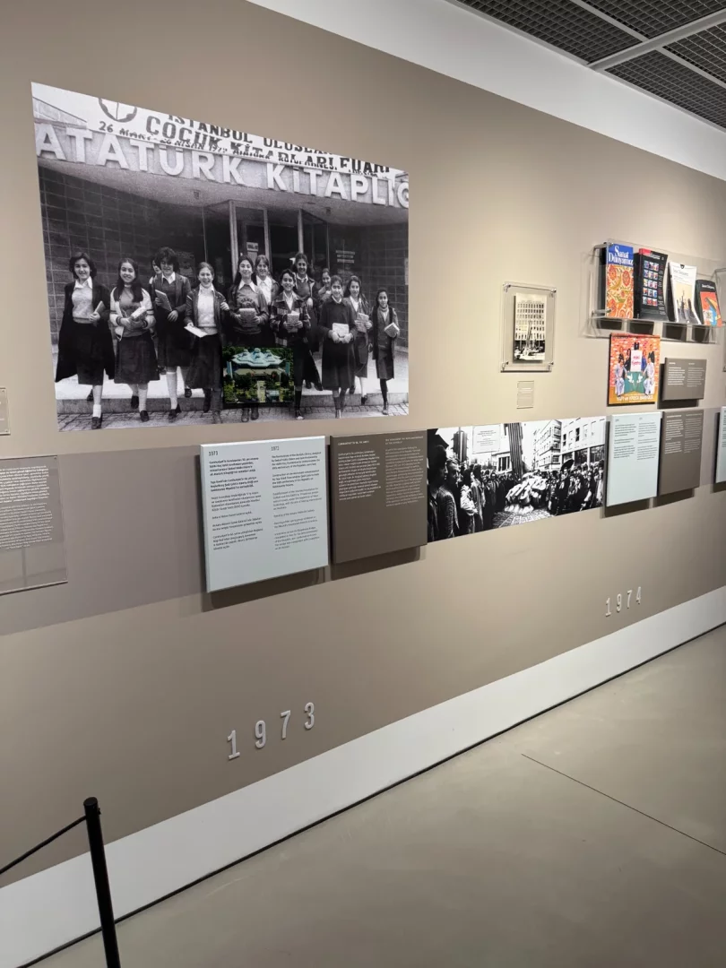 Koç Topluluğu'nun yüzyılın izleri ve sanata katkıları temalı bir sergide, İstanbul Atatürk Kütüphanesi Çocuk Kütüphanesi önünden çıkan neşeli öğrencileri gösteren, üzerine küçük bir detay görseli eklenmiş büyük siyah beyaz bir fotoğraf ve kronolojik bilgi panoları sergileniyor.