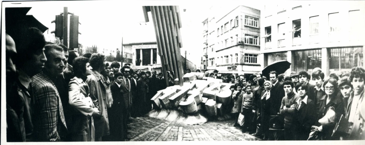 Bu siyah beyaz fotoğrafta, bir şehir meydanında veya caddesinde, dönemin giysileriyle kalabalık bir grup, yere düşmüş ve ayakta duran parçalardan oluşan soyut bir heykelin etrafında toplanmış, Koç Topluluğu tarafından desteklenen bir sanat eserinin açılışını veya sergisini gözlemliyor gibi görünüyor.