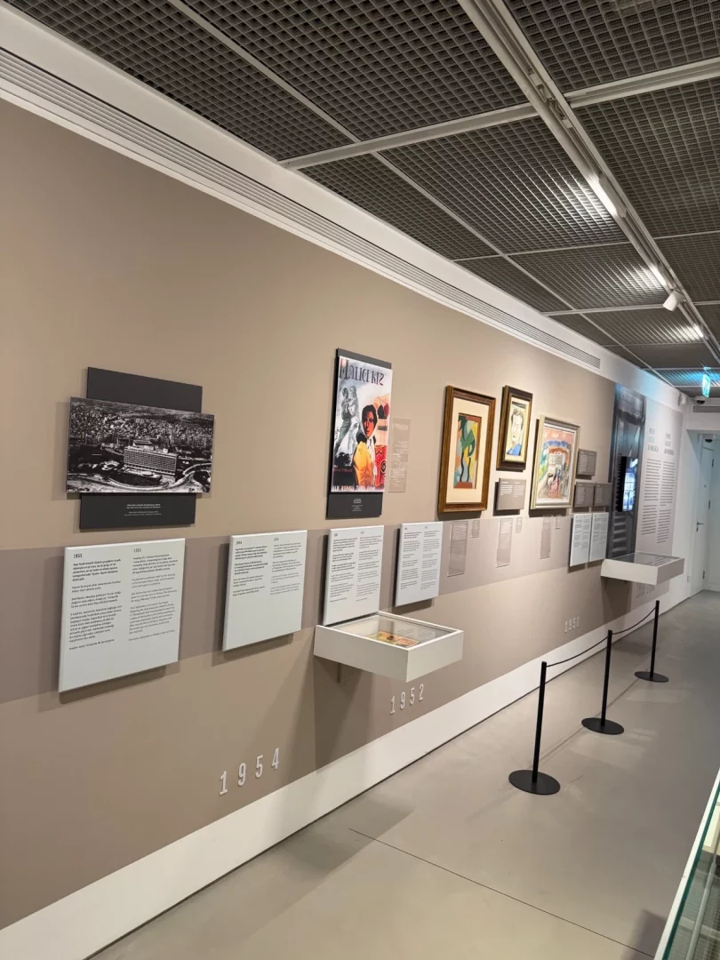Duvar boyunca uzanan, büyük yılların ve bilgi panellerinin eşlik ettiği fotoğraflar, çeşitli sanat eserleri ve vitrinlerdeki küçük objelerle Koç Topluluğu'nun