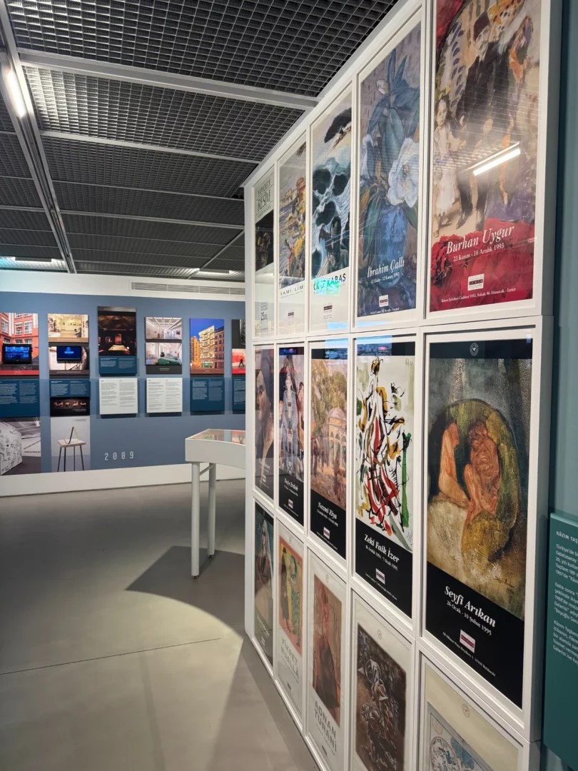 Bir modern sanat sergisi salonunda, Burhan Uygur, İbrahim Çallı ve Seyfi Arıkan gibi önemli sanatçıların çeşitli eserlerini ve sergi bilgilerini içeren çok sayıda çerçeveli posterin sıralandığı uzun bir duvar, Türkiye sanat tarihindeki yüzyılın izlerini yansıtırken, sol tarafta mimari projelerin yer aldığı daha küçük panolar görülmektedir.