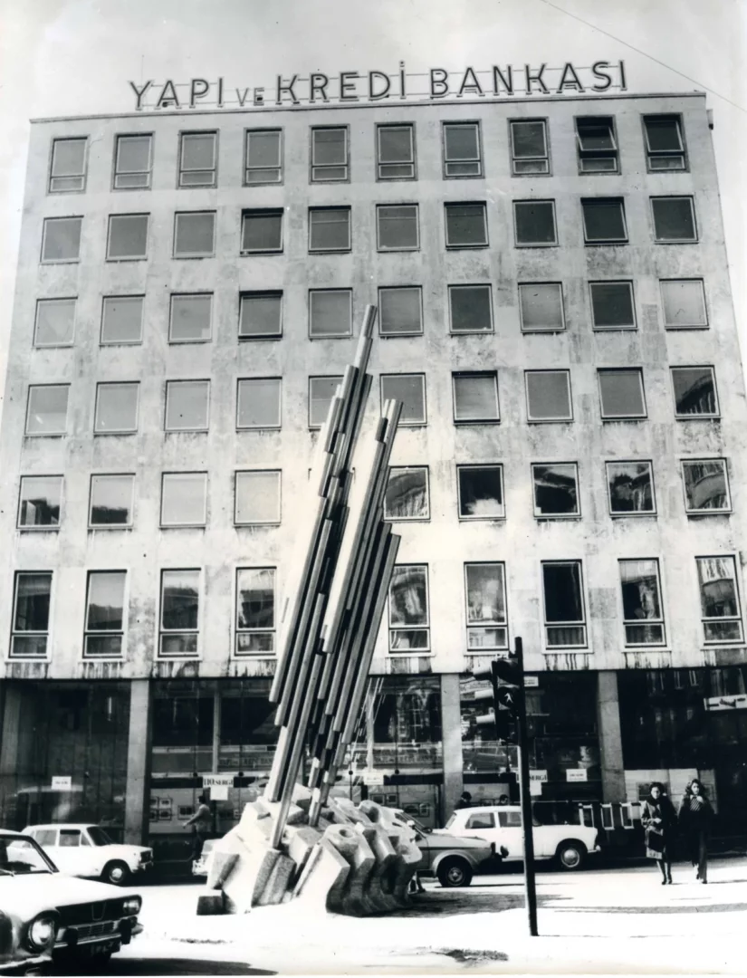 Siyah beyaz bu tarihi fotoğrafta, tepesinde YAPI ve KREDİ BANKASI yazan yüksek bir binanın önünde, Koç Topluluğu'nun sanata verdiği değeri temsil eden soyut bir heykel ve 20. yüzyılın şehir yaşantısını yansıtan hareketli bir cadde ile araçlar ve yayalar görülmektedir.