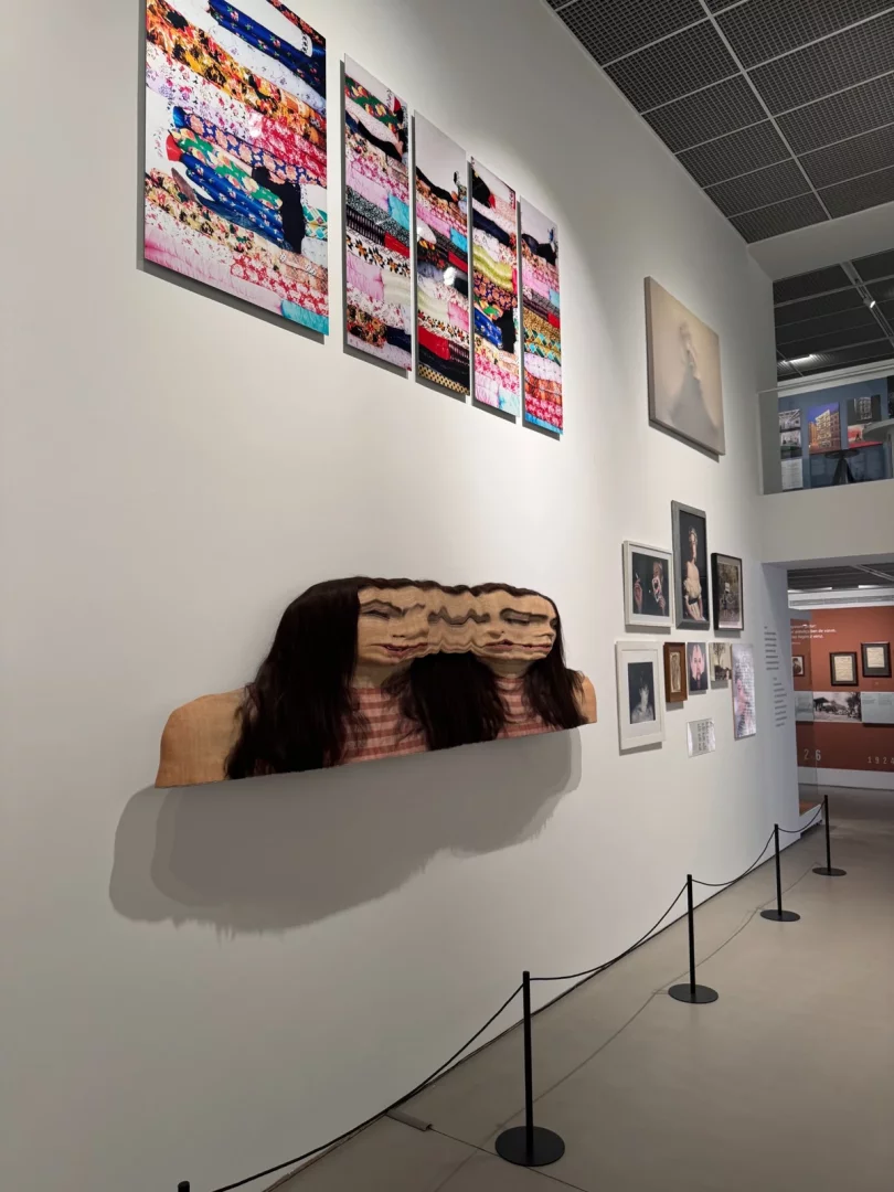 Beyaz bir sanat galerisi duvarında, üstte renkli, katmanlı kumaş desenlerini yansıtan beş dikey panel ve hemen altında dalgalı bir görselle uzun saçlı bir kadın figürünün ahşap dokulu, çağdaş bir büst heykeli ile sağda çeşitli çerçeveli portre ve soyut sanat eserlerinin sergilendiği, Koç Topluluğu ve sanat temasını yansıtan