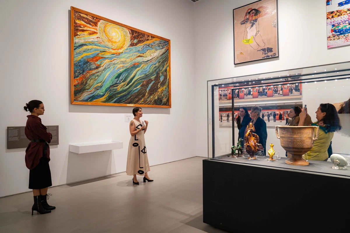 Sanat galerisinde, canlı renklerle boyanmış büyük, soyut bir tabloya hayranlıkla bakan bir ziyaretçi ve şık giyimli bir rehberin açıklamalarını dinleyen diğer misafirler, yüzyılın izlerini taşıyan modern ve tarihî sanat eserleriyle dolu cam vitrin önünde, sanatın büyüleyici atmosferini deneyimliyor.