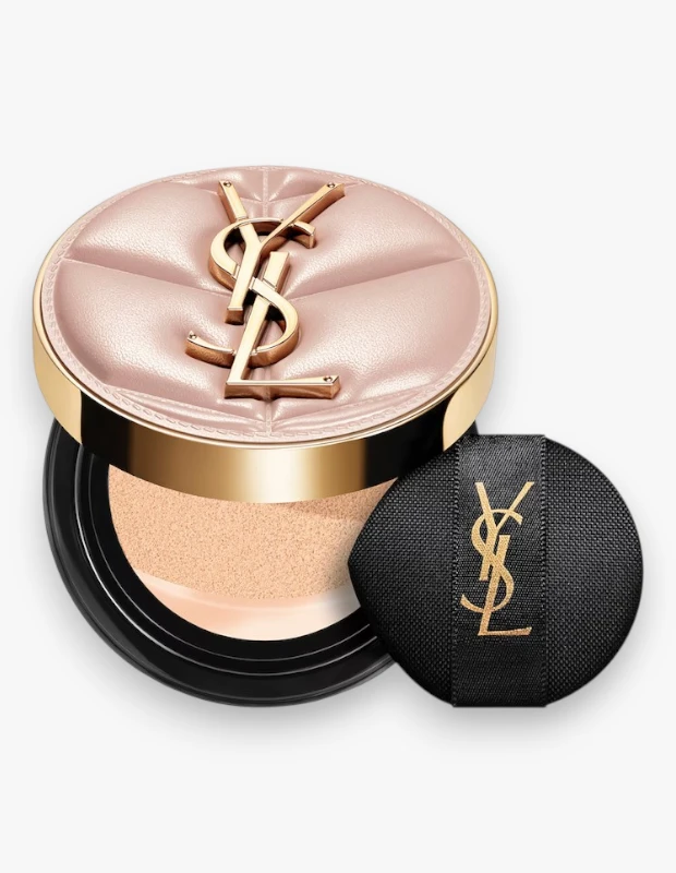 Yves Saint Laurent Skin Affair Cushion Fondöten, kapitone desenli açık pembe kapağında altın rengi YSL logosu taşıyan yuvarlak şık bir kutuda, açık renkli fondöten süngeri görünecek şekilde açılmış ve yanında altın YSL logosu işli siyah makyaj süngeriyle birlikte zarifçe duruyor.
