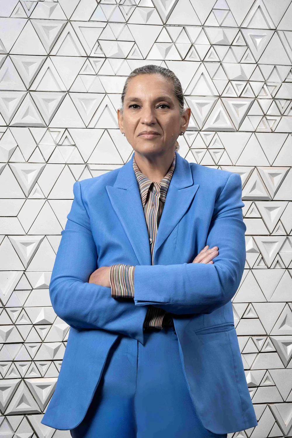 Zeynep Bodur Okyay, arkasında beyaz renkli, üç boyutlu üçgen desenlere sahip modern bir duvarın önünde, canlı mavi bir takım elbise ve çok renkli çizgili bir gömlek giymiş, kolları çapraz bir şekilde kameraya doğrudan bakıyor, bu Artweeks içeriği için çekilmiş bir portre fotoğrafı.